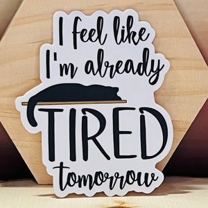 May include: White sticker with black text that reads "I feel like I'm already tired tomorrow". A black cat is sleeping on a black line above the word "tired".