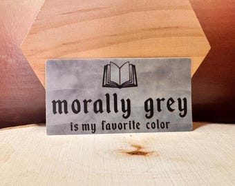 May include: A rectangular, gray sign with the text "morally grey is my favorite color" in black gothic font. An open book graphic is above the text. The sign is set against a wooden background.