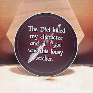 May include: A round sticker with a black background and red text that reads "The DM killed my character and all I got was this lousy sticker." A silver sword is depicted in the background.