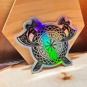 May include: A holographic sticker with a black and white design of two crossed axes and a circle with runes and a symbol in the center. The background of the sticker is iridescent.