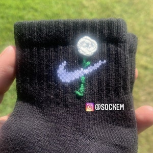 May include: Black socks with a white flower and a blue and green Nike swoosh embroidered on them. The socks have the Instagram handle @sockem on them.