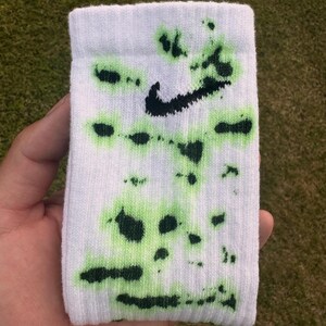 May include: White athletic sock with a black Nike swoosh logo and green splatter design. The sock is held up against a background of green grass.