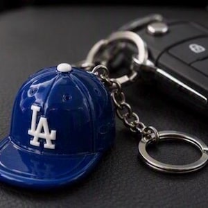 May include: A glossy blue baseball cap keyring featuring the white LA logo on the front and a small white button on top. The cap is attached to a silver metal chain and a circular split ring, positioned next to a black car key fob.