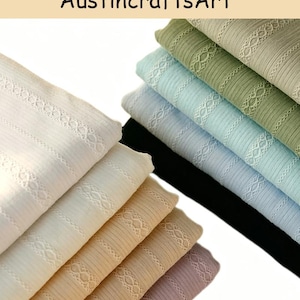 May include: A stack of folded ribbed fabric pieces in various colors including white, beige, light pink, black, light blue, and sage green. Each piece features a vertical decorative lace trim with a repeating diamond pattern.