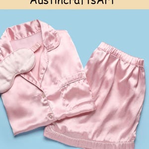 May include: A light pink pajama set featuring a button-down shirt with a collar and a chest pocket, paired with matching elastic-waist shorts and a coordinating sleep mask. The fabric has a smooth, satin-like finish. Text reads AustincraftsArt.