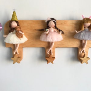 May include: A wooden wall-mounted coat rack with three hooks shaped like stars. Each hook has a felt doll hanging from it. The dolls are wearing dresses and have long hair. The dolls are wearing different colored dresses and have different accessories.