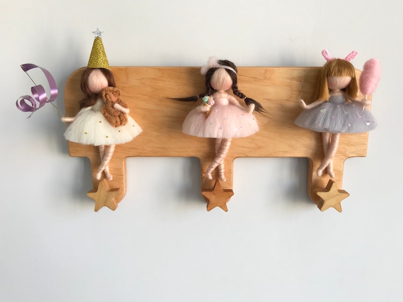 May include: A wooden wall-mounted coat rack with three hooks shaped like stars. Each hook has a felt doll hanging from it. The dolls are wearing dresses and have long hair. The dolls are wearing different colored dresses and have different accessories.