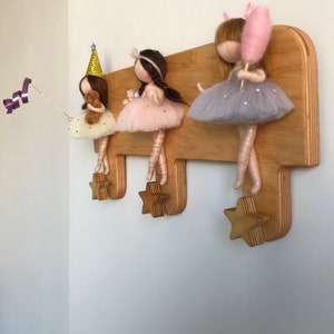 May include: Wooden coat rack with three star-shaped hooks. Three felt dolls are hanging from the hooks. The dolls are wearing dresses and have different accessories, including a party hat, a teddy bear, a gift box, and a pink cotton candy.