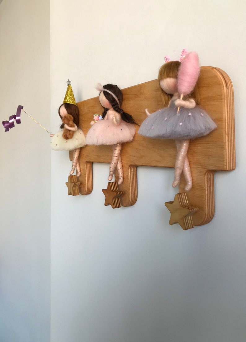 May include: Wooden coat rack with three star-shaped hooks. Three felt dolls are hanging from the hooks. The dolls are wearing dresses and have different accessories, including a party hat, a teddy bear, a gift box, and a pink cotton candy.