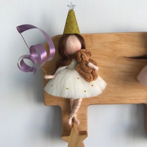 May include: A wooden wall-mounted coat rack with a star-shaped hook. A felt doll with a gold glitter party hat and a brown teddy bear is hanging from the hook. The doll is wearing a white dress with gold glitter stars and a purple ribbon.