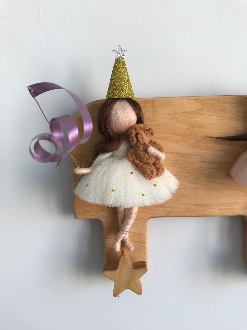 May include: A wooden wall-mounted coat rack with a star-shaped hook. A felt doll with a gold glitter party hat and a brown teddy bear is hanging from the hook. The doll is wearing a white dress with gold glitter stars and a purple ribbon.