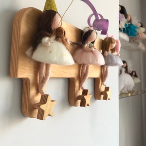 May include: Wooden wall-mounted coat rack with three star-shaped hooks. The rack is decorated with four felt fairy dolls wearing dresses and hats. The fairies are holding a small teddy bear, a cupcake, and a flower.
