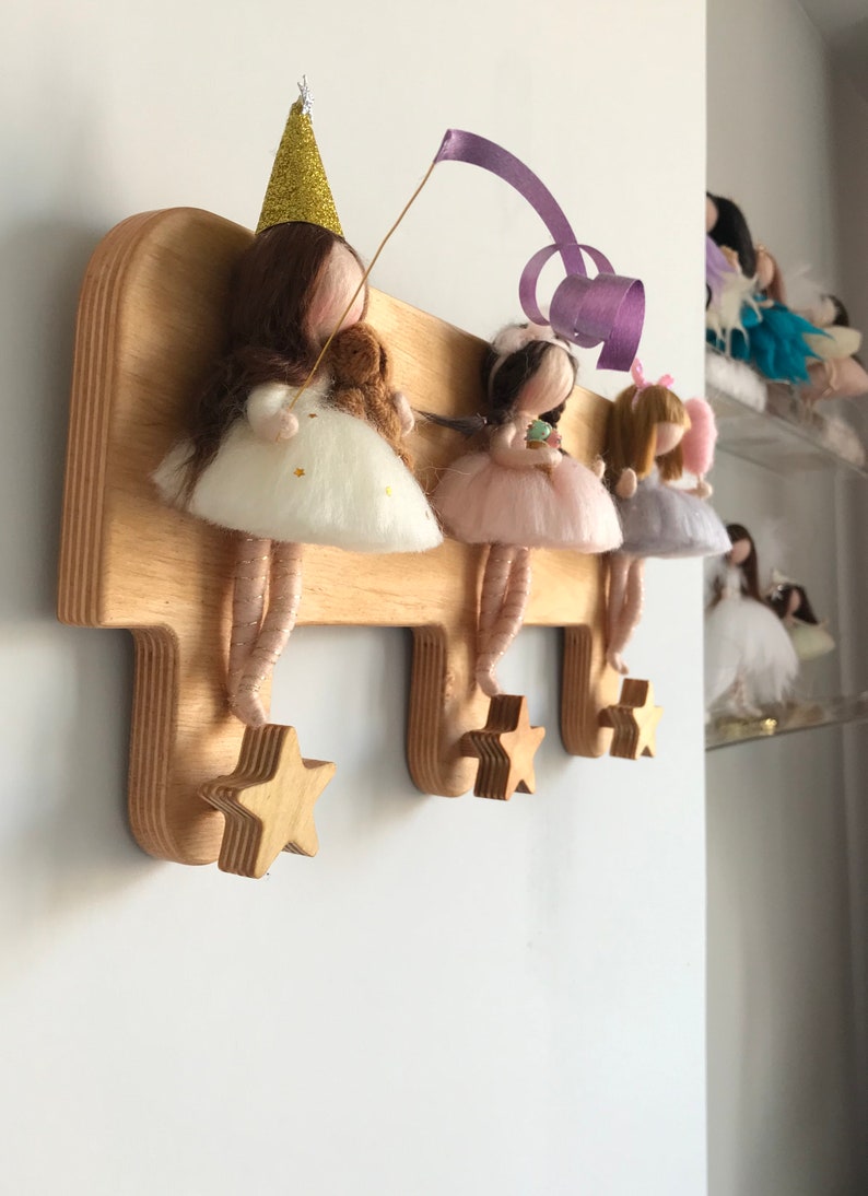 May include: Wooden wall-mounted coat rack with three star-shaped hooks. The rack is decorated with four felt fairy dolls wearing dresses and hats. The fairies are holding a small teddy bear, a cupcake, and a flower.