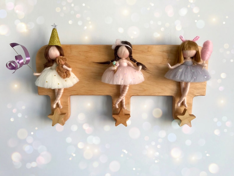 May include: Three felt doll figures with long hair and dresses hanging on a wooden wall hook with star-shaped hooks. The dolls are wearing a white dress with gold glitter, a pink dress with glitter, and a gray dress with glitter.