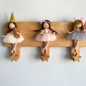 May include: Wooden wall-mounted coat rack with three star-shaped hooks. Each hook features a felt doll with a different dress and accessories. The dolls are wearing pink, white, and grey dresses with glitter accents.