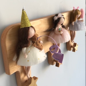 May include: Wooden wall-mounted coat rack with three hooks shaped like stars. Each hook has a felt doll hanging from it. The dolls are dressed in different colors and have different accessories.