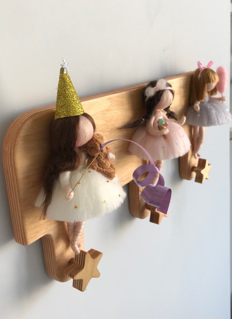 May include: Wooden wall-mounted coat rack with three hooks shaped like stars. Each hook has a felt doll hanging from it. The dolls are dressed in different colors and have different accessories.