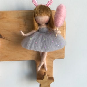 May include: A wooden doll hanger with a star-shaped hook. A felt doll with blonde hair, a grey dress, and pink bunny ears is holding a pink cotton candy.