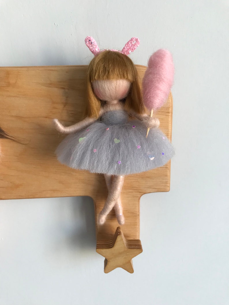 May include: A wooden doll hanger with a star-shaped hook. A felt doll with blonde hair, a grey dress, and pink bunny ears is holding a pink cotton candy.