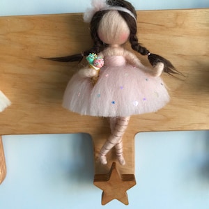 May include: A felt doll with brown hair, wearing a pink dress with sequins and holding an ice cream cone. The doll is attached to a wooden star-shaped hook.