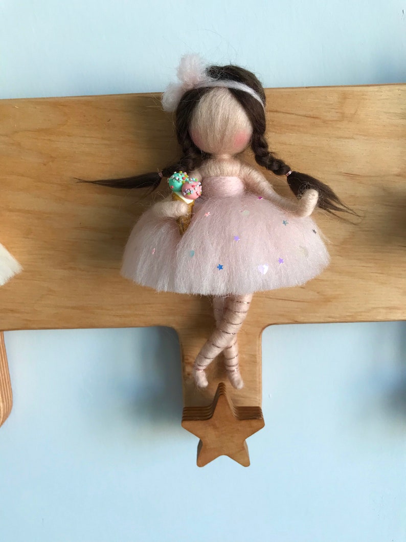 May include: A felt doll with brown hair, wearing a pink dress with sequins and holding an ice cream cone. The doll is attached to a wooden star-shaped hook.
