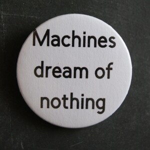 May include: A circular white pin-back button featuring the black text Machines dream of nothing printed in a simple sans-serif font. The button has a smooth, matte finish and a clean, minimalist design that is centred on the white surface.