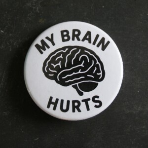 May include: A circular white pin-back button featuring a black graphic illustration of a human brain in the center. The text "MY BRAIN" is printed in black capital letters above the brain, and the word "HURTS" is printed below it.