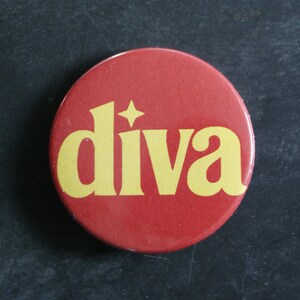 May include: A circular pinback button featuring a solid red background with the word diva printed in a bold yellow serif font. The letter i is topped with a four-pointed star shape instead of a dot, centered on the face of the round button.