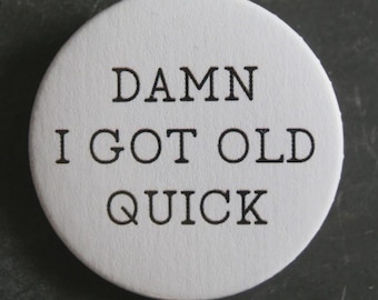 May include: A white, circular button with the phrase "DAMN I GOT OLD QUICK" printed in black, sans-serif font. The button has a slightly textured surface and a simple, straightforward design.