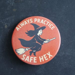 May include: A round, orange pin-back button featuring a black illustration of a witch flying on a broomstick. The text "ALWAYS PRACTICE" is printed in white along the top curve, and "SAFE HEX" is printed in white along the bottom curve.