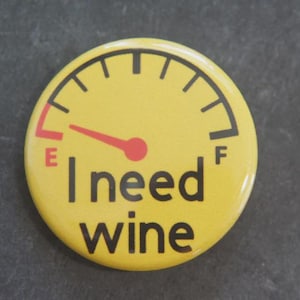 May include: A round yellow pinback button featuring a black fuel gauge graphic with a red needle pointing towards the E. The text I need wine is printed in black below the gauge, with the letters E and F marking the empty and full levels.