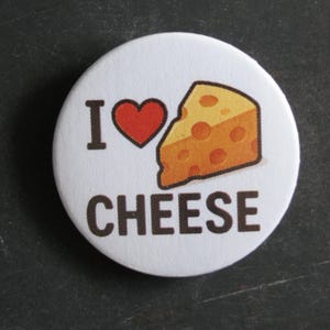 May include: A circular white pin-back button featuring the black text I, a red heart symbol, a yellow wedge of Swiss cheese with holes, and the black text CHEESE centred below the graphic. The design is printed on a smooth, round surface.
