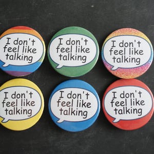 May include: Six circular pin-back badges arranged in two rows. Each badge features a white speech bubble containing the black text 'I don't feel like talking'. The backgrounds include rainbow stripes, solid green, glittery pink, solid yellow, solid blue, and solid red.
