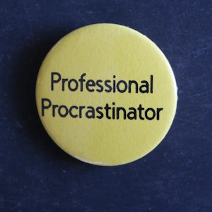 May include: A circular yellow pin-back button featuring the black text Professional Procrastinator printed in a sans-serif font. The button has a smooth, matte finish and a simple, clean design with the text centred on the yellow background.
