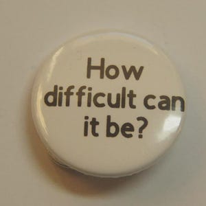 May include: A circular white pin-back button featuring the text "How difficult can it be?" printed in a dark gray, sans-serif font. The button has a smooth, glossy finish and a standard round shape, presenting a clear and legible message on its surface.
