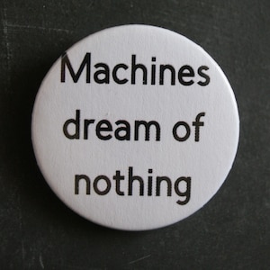 May include: A circular white pin-back badge featuring the black text "Machines dream of nothing" printed in a clean, sans-serif font. The badge has a smooth, matte finish and a simple, minimalist design that centres the text on the white surface.
