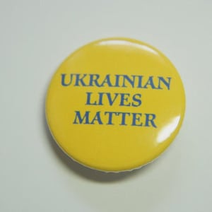 May include: A circular yellow pin-back button featuring the text UKRAINIAN LIVES MATTER printed in blue capital letters. The button has a smooth, glossy finish and a simple, clean design that highlights the bold blue lettering against the yellow background.