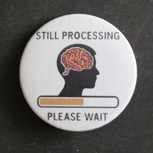 Könnte beinhalten: Ein runder weißer Button mit der schwarzen Silhouette eines menschlichen Kopfes und einer rosa Gehirngrafik darin. Unter dem Kopf befindet sich ein horizontaler Ladebalken, der teilweise beige gefüllt ist. Der Text STILL PROCESSING und PLEASE WAIT.