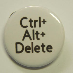 May include: A round white pin-back button featuring the black text Ctrl+ Alt+ Delete printed in a sans-serif font. The circular accessory has a smooth, glossy surface and a simple design that displays the common computer keyboard command.