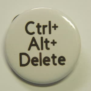 May include: A round white pin-back button featuring the black text Ctrl+ Alt+ Delete printed in a sans-serif font. The circular accessory has a smooth, glossy surface and a simple design that displays the common computer keyboard command.