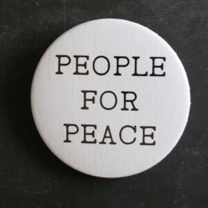 May include: A circular white button featuring the black text PEOPLE FOR PEACE printed in a clean, sans-serif font. The design is centred on the round surface, providing a clear and legible message against the solid white background of the pin.