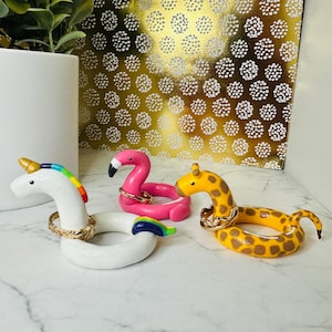 Könnte beinhalten: Drei skurrile Ringhalter in Form eines Einhorns, eines Flamingos und einer Giraffe. Das Einhorn ist weiß mit einer Regenbogenmähne und einem Regenbogen-Schweif. Der Flamingo ist rosa und die Giraffe ist gelb mit braunen Flecken. Jedes Tier hat einen goldfarbenen Ring um den Hals.