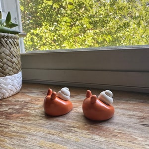 May include: Two small ceramic snails with orange bodies and white shells. The snails are sitting on a wooden surface.