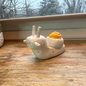 Könnte beinhalten: Eine weiße Keramik-Schneckenfigur mit schwarzen Punkten, einer gelben und orangen Schale und einer glänzenden Oberfläche.