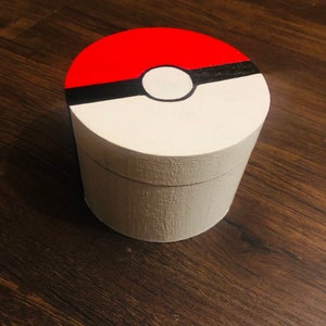 May include: A white cylindrical wooden box with a red, black, and white Pokeball design painted on the lid.