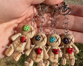 May include: Five handmade voodoo doll keychains with different colored buttons for eyes and red hearts. The dolls are made of beige clay and have a stitched X across their bodies. The keychains are attached to silver chains.