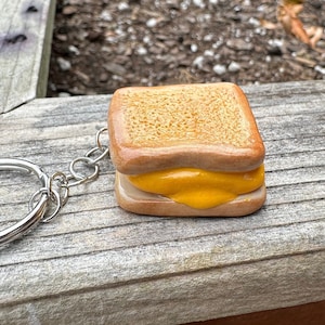 May include: A small, brown, ceramic keychain shaped like a grilled cheese sandwich. The keychain has a metal ring attached to it.