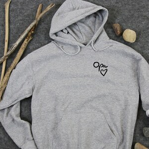 Peut inclure: Un sweat à capuche gris avec un motif brodé noir qui dit "Opa" avec un cœur.