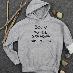 Peut inclure: Sweat-shirt à capuche gris avec le texte "SOON TO BE GRANDPA" et une flèche avec un cœur au milieu.