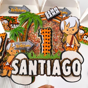 Puede incluir: Un topper de pastel con temática de Los Picapiedra con una palmera verde, un número uno con estampado de animal naranja y negro, un personaje de dibujos animados y el nombre Santiago. El topper está decorado con chispas naranja y blancas.